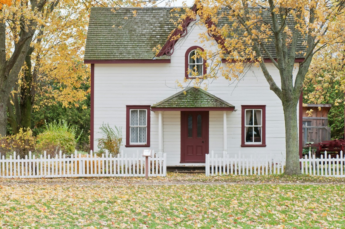 Picture of a cute white house with a white picket fence in the fall with yellow leaves dropping onto a lawn in front